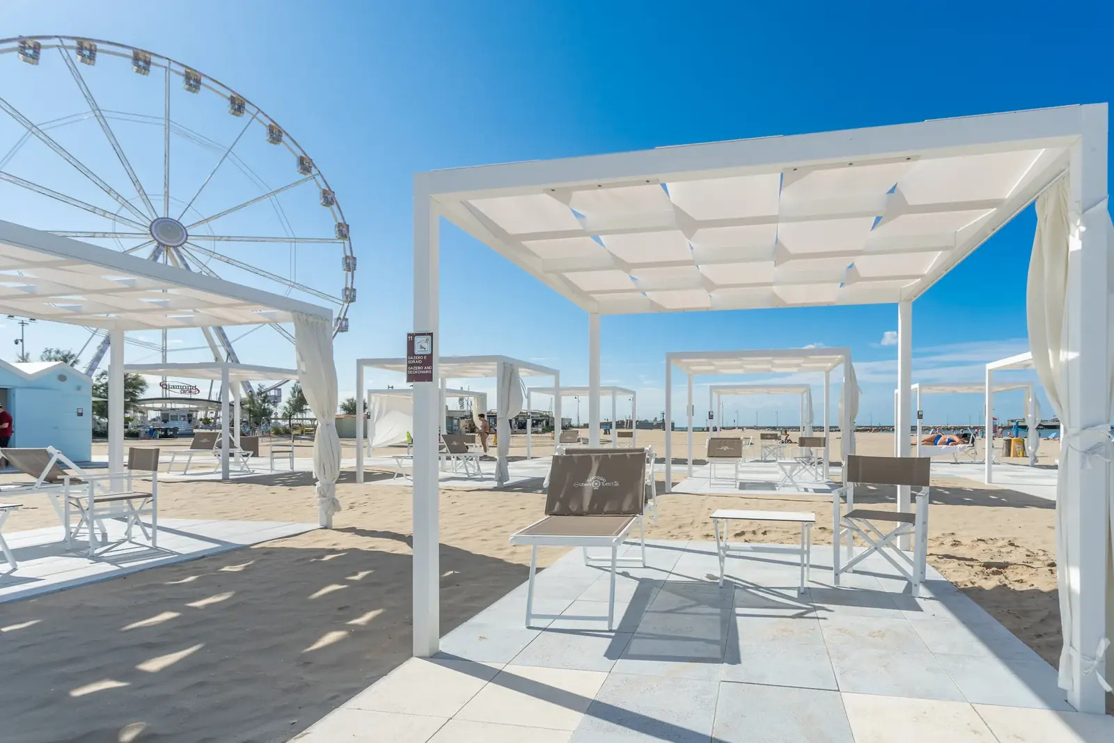 Le pergole bianche della spiaggia libera attrezzata di Rimini, con le sedute sulla sabbia e la ruota panoramica sullo sfondo