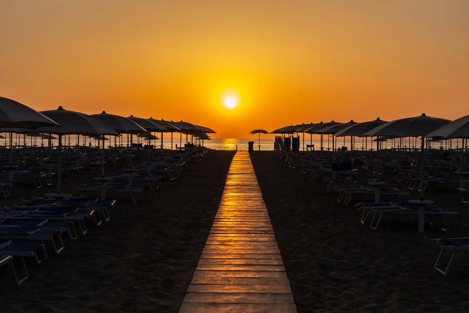 Una passerella di legno fra due file di ombrelloni chiusi sulla spiaggia di Rimini, con il sole che sorge sul mare
